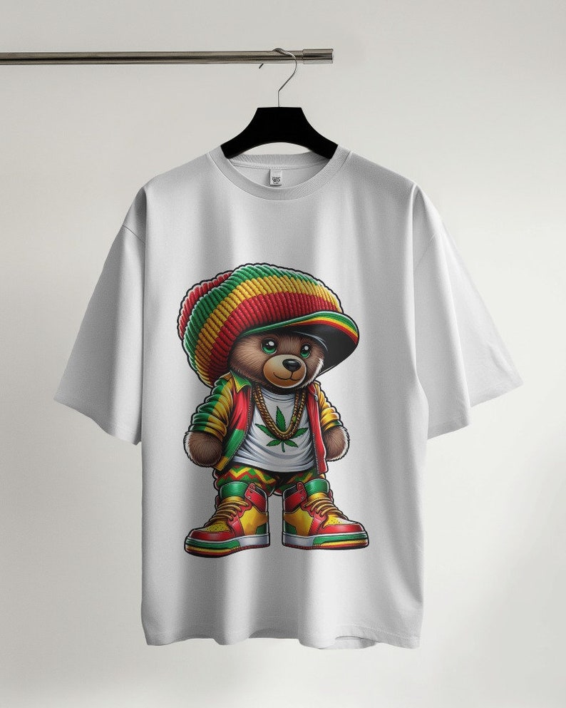 Puede incluir: Camiseta blanca con un gr&aacute;fico de un oso de peluche de dibujos animados que lleva un sombrero rojo, amarillo y verde, una cadena de oro y un dise&ntilde;o de hoja verde. El oso lleva ropa roja, amarilla y verde y zapatillas rojas y amarillas.