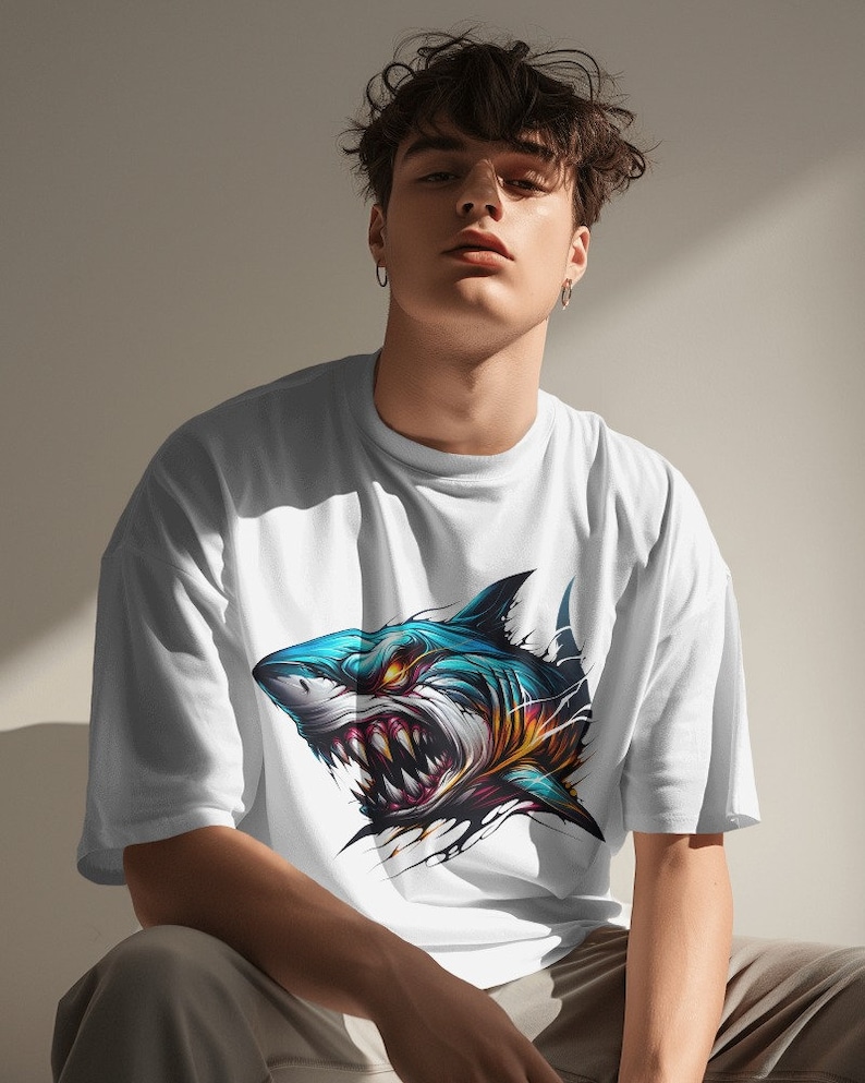 May include: A white t-shirt with a graphic print of a blue and yellow shark with sharp teeth. The shark is shown with its mouth open and its eyes are glowing red.