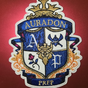 May include: An embroidered patch featuring a shield crest with a gold crown at the top. The shield is divided into four sections with a blue letter A, a blue helmet, a pink rose, and a blue letter P. A gold lion head sits in the center. The text AURADON PREP is visible.