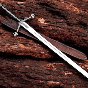 May include: A silver and steel broadsword with a decorative hilt and crossguard, resting in a brown leather sheath. The sword is set against a dark, textured wooden background.