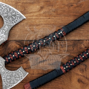 May include: Two Viking axes with intricately etched silver blades and wooden handles wrapped in black and red leather. The axes feature a compass design and wolf imagery. The handles are adorned with decorative studs.