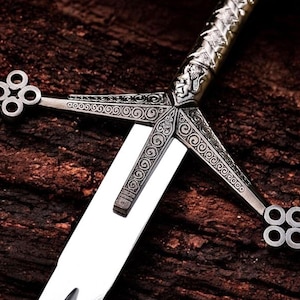 May include: A detailed close-up of a silver sword with an ornate handle and crossguard. The blade is bright and polished, contrasting with the darker, textured wooden background. The crossguard features intricate scrollwork and a quatrefoil design.