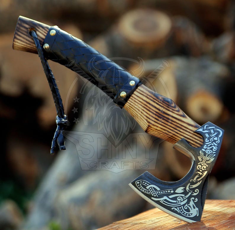 May include: A small black-handled axe with a wooden handle and a black leather wrap. The axe head has a detailed design with intricate patterns and a small hole at the top. The axe is resting on a wooden surface.
