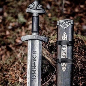 May include: A detailed image of a Viking sword and scabbard. The sword features a silver blade with the word "NORTHMEN" etched into it. The handle is dark wood with silver accents. The scabbard is also dark wood with silver detailing and black leather straps.