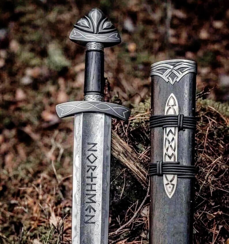 May include: A detailed image of a Viking sword and scabbard. The sword features a silver blade with the word "NORTHMEN" etched into it. The handle is dark wood with silver accents. The scabbard is also dark wood with silver detailing and black leather straps.