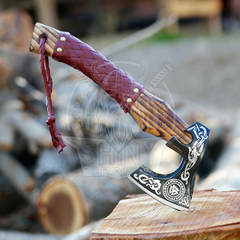 May include: A black and silver axe with a wooden handle wrapped in red leather. The axe head has a Viking symbol engraved on it. The axe is resting on a piece of wood.