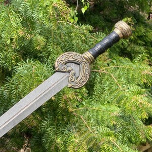 May include: A decorative sword with a patterned blade and ornate handle. The handle features a black grip, gold accents, and a detailed circular guard. The sword is set against a backdrop of green foliage.