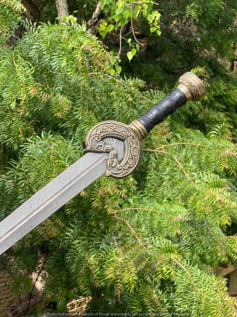 May include: A decorative sword with a patterned blade and ornate handle. The handle features a black grip, gold accents, and a detailed circular guard. The sword is set against a backdrop of green foliage.