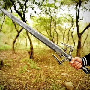 May include: A detailed, silver-colored sword with an ornate handle and a textured blade. The sword features a unique design with three curved prongs extending from the handle. The background is a blurred forest scene.