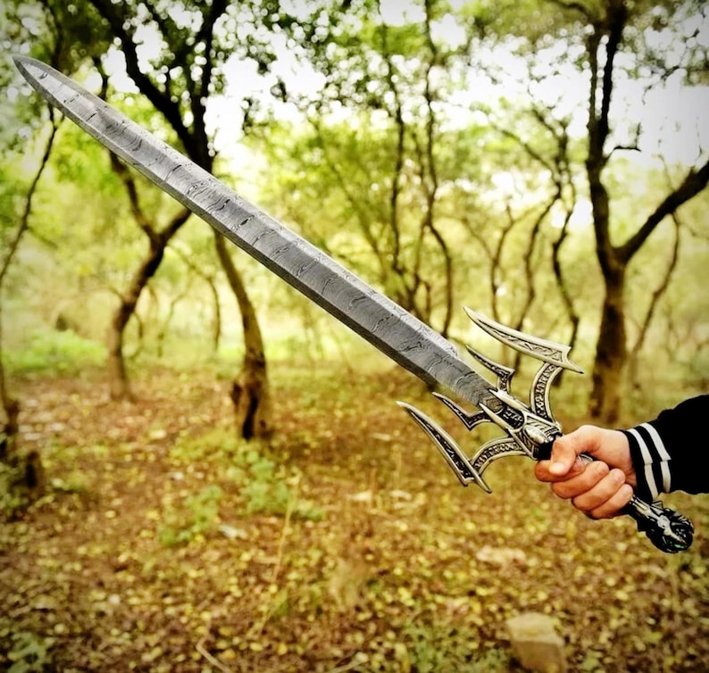 May include: A detailed, silver-colored sword with an ornate handle and a textured blade. The sword features a unique design with three curved prongs extending from the handle. The background is a blurred forest scene.