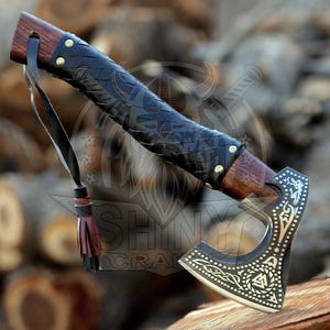 May include: A black and silver Viking axe with a leather wrapped handle and a decorative brass design. The axe has a tassel on the end of the handle.
