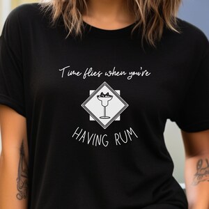 May include: Black t-shirt with white text that reads "Time flies when you're having rum". A white silhouette of a cocktail glass with a lime wedge is inside a white diamond shape.