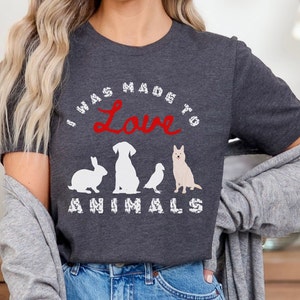 May include: A dark gray t-shirt with the text "I was made to love animals" in white with a red heart. The text is surrounded by a white circle. The shirt features a white silhouette of a rabbit, dog, bird, and dog.