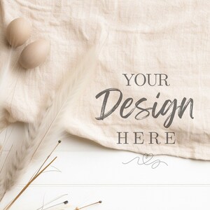 Op de afbeelding: Een platte lay-out mockup met een beige linnen stof gedrapeerd over een wit houten oppervlak. Twee beige eieren liggen op de stof, en een boeket gedroogd pampasgras bevindt zich aan de linkerkant van de afbeelding. De tekst "YOUR Design HERE" is in zwart op de stof geschreven.