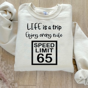 Könnte beinhalten: Ein cremefarbener Pullover mit dem Text "Life is a trip enjoy every mile" und einem schwarz-weißen Geschwindigkeitsbegrenzungszeichen mit der Aufschrift "Speed Limit 65".