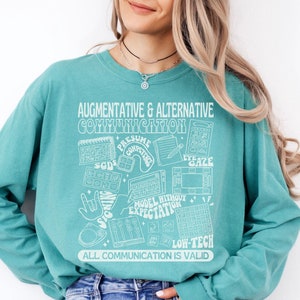 May include: A teal long-sleeve shirt with a white graphic design featuring the text "Augmentative & Alternative Communication" and various symbols related to communication, including sign language, eye gaze, and low-tech communication methods.