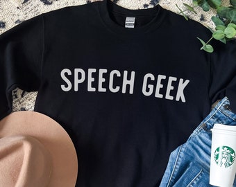 Puede incluir: Sudadera negra con el texto "SPEECH GEEK" en letras blancas. La sudadera está sobre una superficie estampada con un sombrero marrón, unos vaqueros azules y una taza de café blanca con un logotipo de Starbucks verde.