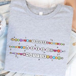 Könnte beinhalten: Graues T-Shirt mit dem Text "YOUR WORDS MATTER" in einer bunten, verspielten Schriftart. Der Text ist von kleinen, bunten Symbolen wie Herzen, Friedenstauben und Smileys umgeben.