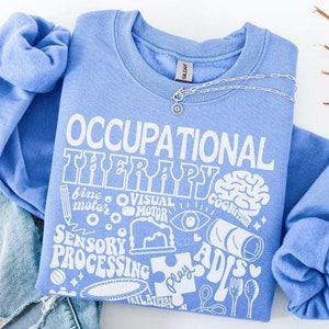 Peut inclure: Un sweat-shirt bleu clair avec le texte "Occupational Therapy" en lettres blanches. Le sweat-shirt présente une variété d'illustrations blanches liées à l'ergothérapie, notamment un cerveau, une main, un puzzle et un crayon.
