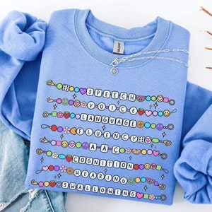 Może przedstawiać: Jasnoniebieska bluza z nadrukiem kolorowych bransoletek, które tworzą napis "SPEECH, VOICE, LANGUAGE, FLUENCY, AAC, COGNITION, HEARING, SWALLOWING". Pod nadrukiem znajduje się napis "CAROLINA BLUE".