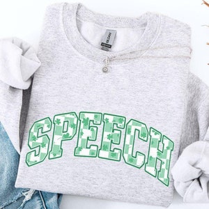 Puede incluir: Una sudadera gris con la palabra "SPEECH" impresa en letras blancas con un patrón de trébol verde. La sudadera está sobre un fondo de mezclilla azul.