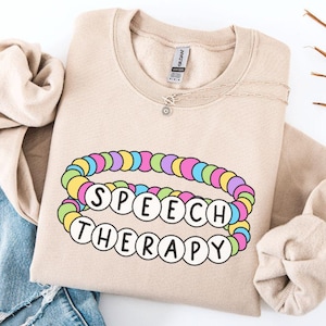 May include: A beige sweatshirt with the words "SPEECH THERAPY" in white letters surrounded by a colorful beaded bracelet design. The sweatshirt is on a white background with blue jeans and a wooden branch.