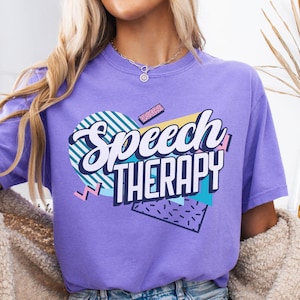 May include: A purple t-shirt with the words "Speech Therapy" in a retro 80s style design. The design includes a heart shape with stripes, a pink rectangle, and a teal triangle with black dots.