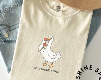 May include: Cream-colored t-shirt with a cartoon goose holding a red telephone. The text "PROFESSIONAL YAPPER" is printed below the goose. The shirt is folded on a light blue denim surface.