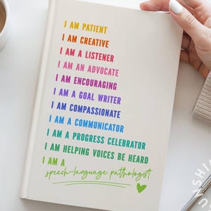 May include: A white journal with colorful text listing positive affirmations. The text includes phrases like "I am patient" and "I am creative." The journal is held by a person, with a pen and a cup of coffee in the background.