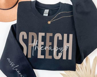 Puede incluir: Sudadera negra con las palabras "SPEECH therapy" impresas en marrón y beige en la parte delantera. Las palabras "miss Laura" están escritas en cursiva en la manga izquierda.