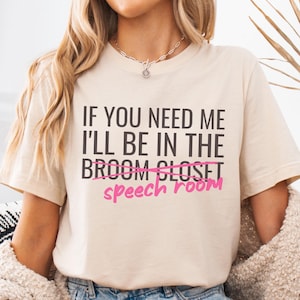 May include: A beige t-shirt with the text "If you need me I'll be in the broom closet speech room" printed on it. The word "closet" is crossed out and "speech room" is written in pink.