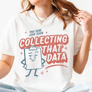 Può includere: T-shirt bianca con la scritta "YOU SURE LOOK GOOD COLLECTING THAT DATA" in rosso e blu. La grafica include un personaggio a fumetti a forma di appunti. La maglietta è in stile casual.