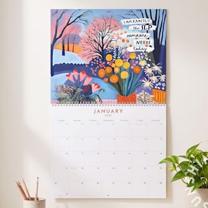 May include: A wall calendar for January 2027 featuring a colorful illustration of a winter landscape with trees, flowers, and the text I AM EXACTLY the SLP someone NEEDS today. The calendar grid is white with black text and a spiral binding at the top.