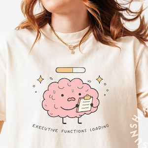 May include: A cream-coloured t-shirt with a cartoon brain holding a clipboard, the text "EXECUTIVE FUNCTIONS LOADING", and a loading bar graphic. The design includes stars and a smiling brain. The shirt is a casual, unisex style.