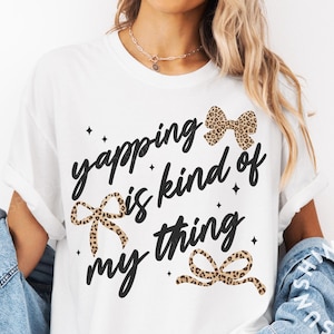 Puede incluir: Camiseta blanca con la frase "yapping is kind of my thing" en escritura negra, adornada con lazos de estampado de leopardo y detalles de estrellas. Una camiseta casual y con estilo.