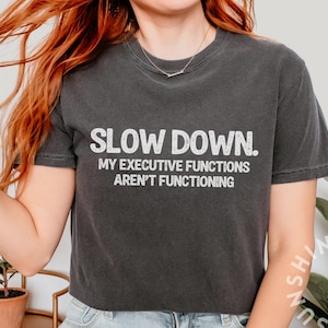 Puede incluir: Camiseta gris oscuro con el texto blanco "SLOW DOWN. MY EXECUTIVE FUNCTIONS AREN'T FUNCTIONING". La camiseta es de cuello redondo y tiene mangas cortas. La camiseta es de estilo vintage.