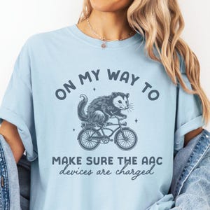 May include: A light blue t-shirt with a graphic of a possum riding a bicycle. The text on the shirt reads "On my way to make sure the AAC devices are charged."