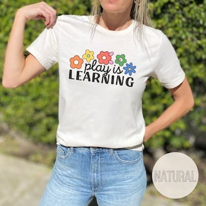 Woman wearing t-shirt that says "Play is Learning" and has cute retro flowers on it for preschool teacher, play therapist, or speech therapist. Gift for teachers or therapists.