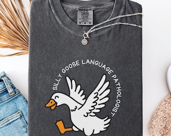 May include: Dark gray Comfort Colors t-shirt with a white goose graphic and the text "SILLY GOOSE LANGUAGE PATHOLOGIST" in a circle. A silver necklace with a sun pendant is on the shirt.