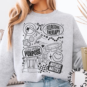 Könnte beinhalten: Hellgraues Sweatshirt mit einem schwarz-weißen Grafikdesign. Das Design enthält die Wörter "Occupational Therapy", "Play with Purpose" und "Movement Matters" sowie Illustrationen einer Schaukel und Blöcken.