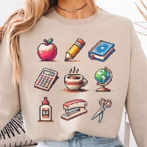 May include: A beige sweatshirt with a pixelated design of school supplies, including an apple, pencil, book, calculator, coffee cup, globe, stapler, glue, and scissors.