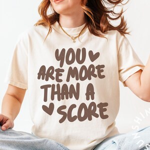May include: Cream-colored t-shirt with the text "YOU ARE MORE THAN A SCORE" in brown letters. The shirt has a relaxed fit and short sleeves. The overall style is casual and comfortable.