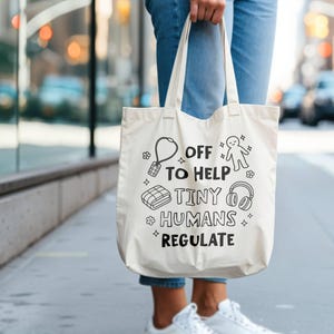 May include: A cream tote bag with black illustrations and text reading "OFF TO HELP TINY HUMANS REGULATE." The design includes a key, person, blanket, and headphones. The bag is held by a person wearing blue jeans.