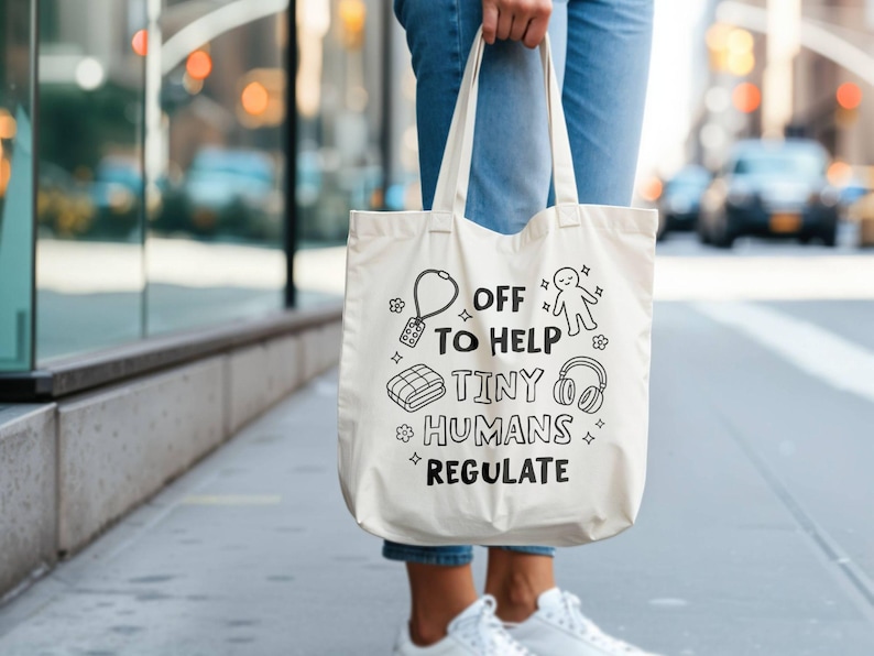 May include: A cream tote bag with black illustrations and text reading "OFF TO HELP TINY HUMANS REGULATE." The design includes a key, person, blanket, and headphones. The bag is held by a person wearing blue jeans.