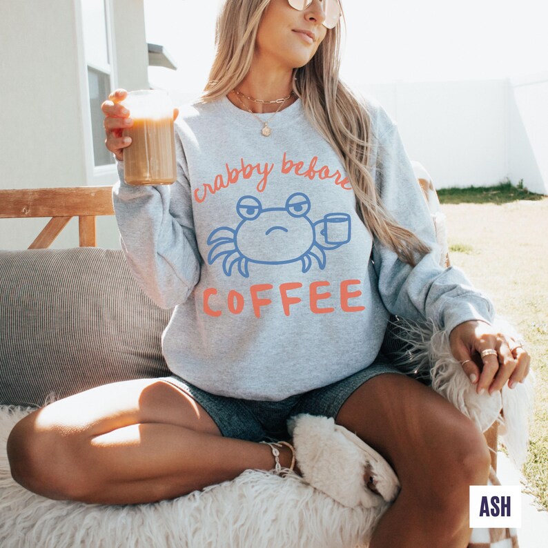 以下が含まれることがあります： 「crabby before coffee」の文字とカニのイラストがプリントされたライトグレーのスウェットシャツ。青いカニがコーヒーカップを持っています。「coffee」の文字はオレンジ色です。カジュアルなスタイルのスウェットシャツです。