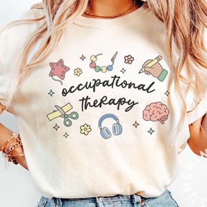 Peut inclure: T-shirt crème avec l'inscription "occupational therapy" en écriture noire. Le t-shirt présente des illustrations colorées d'une étoile, de ciseaux, d'un casque, d'un cerveau et d'autres icônes liées à la thérapie. Le motif est entouré de petites étoiles et de fleurs.
