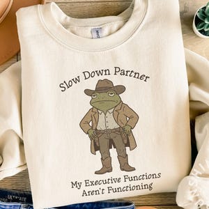 May include: Cream-colored sweatshirt with a cartoon frog dressed as a cowboy. The frog wears a brown hat, coat, and boots, with the text "Slow Down Partner" and "My Executive Functions Aren't Functioning".