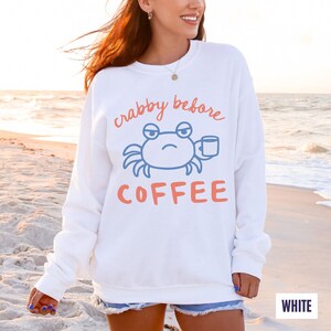 以下が含まれることがあります： 「crabby before COFFEE」の文字と、コーヒーカップを持った青いカニのグラフィックがプリントされた白いスウェットシャツ。テキストとカニはオレンジと青の色合いです。スウェットシャツはビーチで着用されています。
