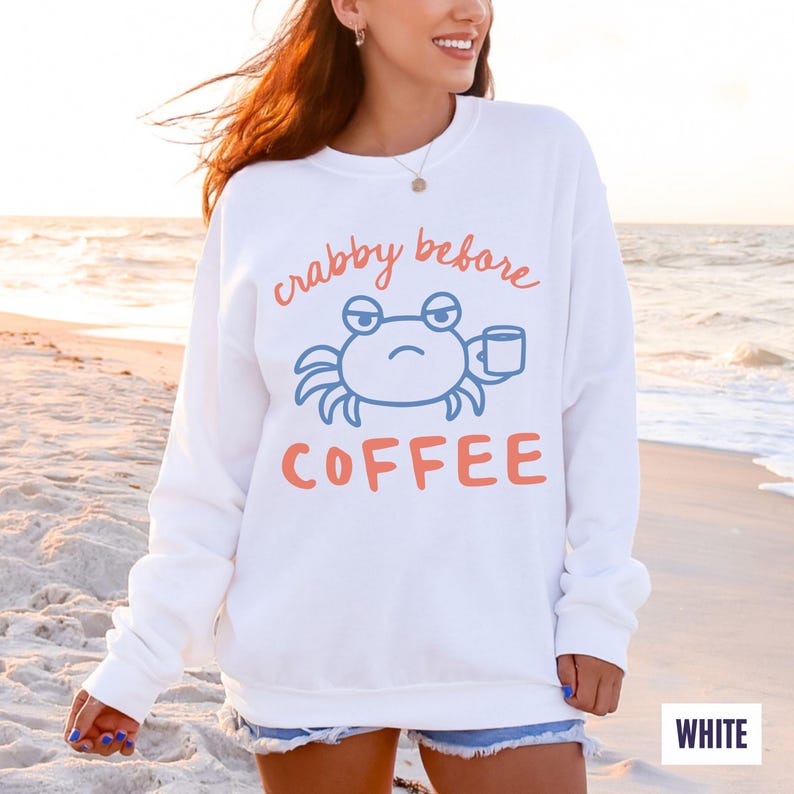 以下が含まれることがあります： 「crabby before COFFEE」の文字と、コーヒーカップを持った青いカニのグラフィックがプリントされた白いスウェットシャツ。テキストとカニはオレンジと青の色合いです。スウェットシャツはビーチで着用されています。