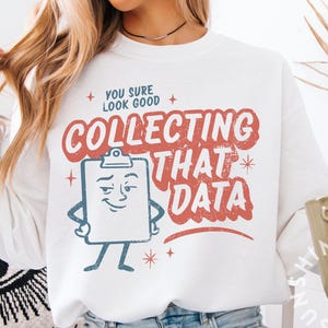 Könnte beinhalten: Weißes Langarm-Sweatshirt mit dem Text "YOU SURE LOOK GOOD COLLECTING THAT DATA" in Rot und Weiß. Die Grafik zeigt eine Zeichentrickfigur eines Klemmbretts. Eine Kaffeetasse ist in der unteren rechten Ecke sichtbar.